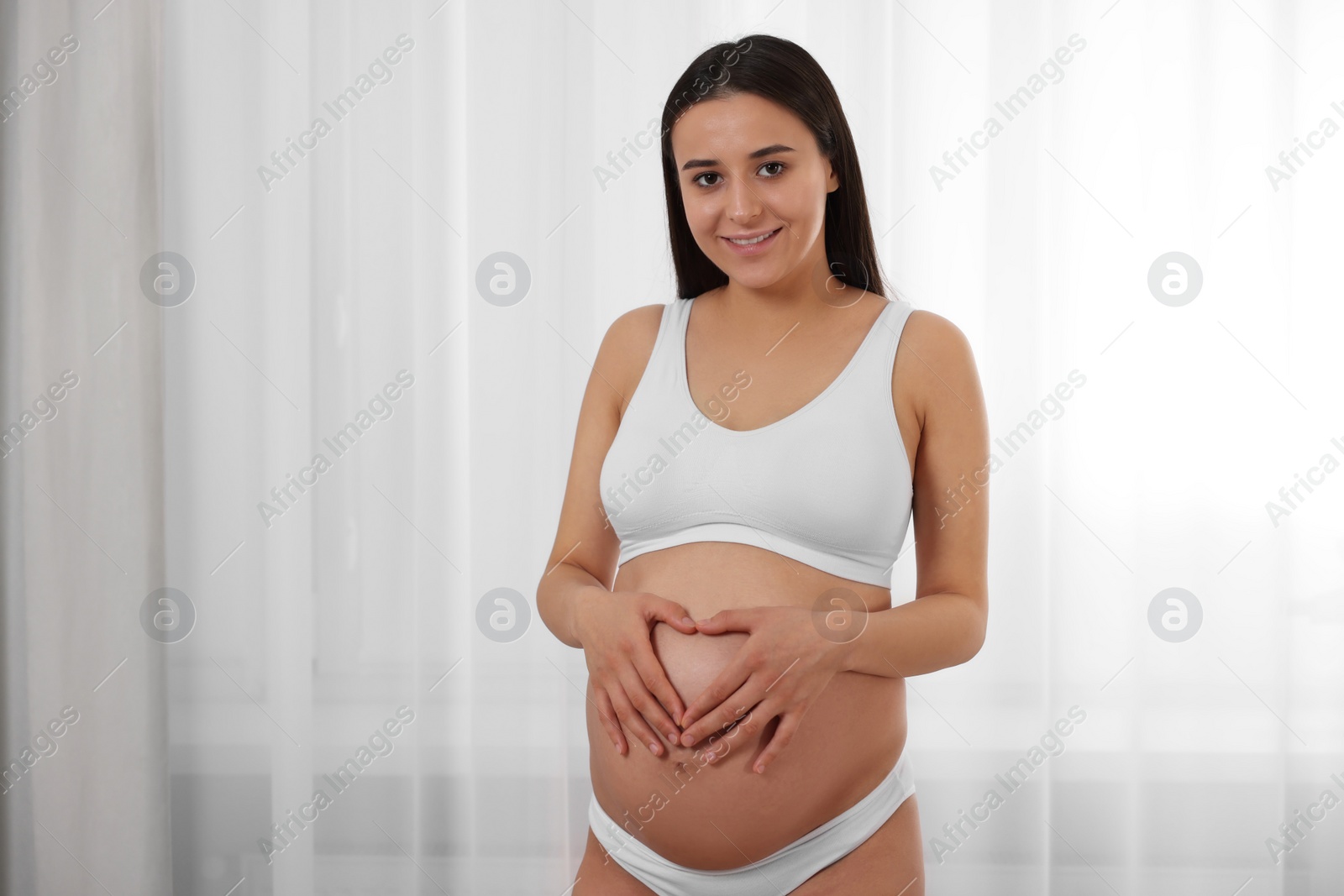 Photo of Beautiful pregnant woman in stylish comfortable underwear making heart with hands on her belly indoors