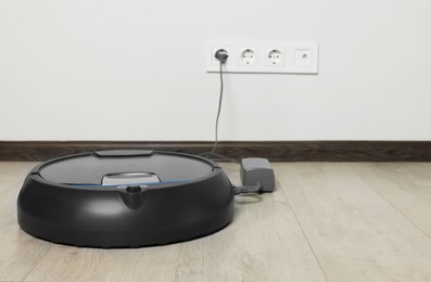 Robotic vacuum cleaner charging on wooden floor near white wall, space for text