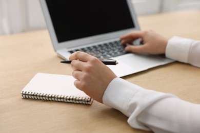 Photo of Woman with notebook and pen working on laptop at wooden table, closeup. Electronic document management