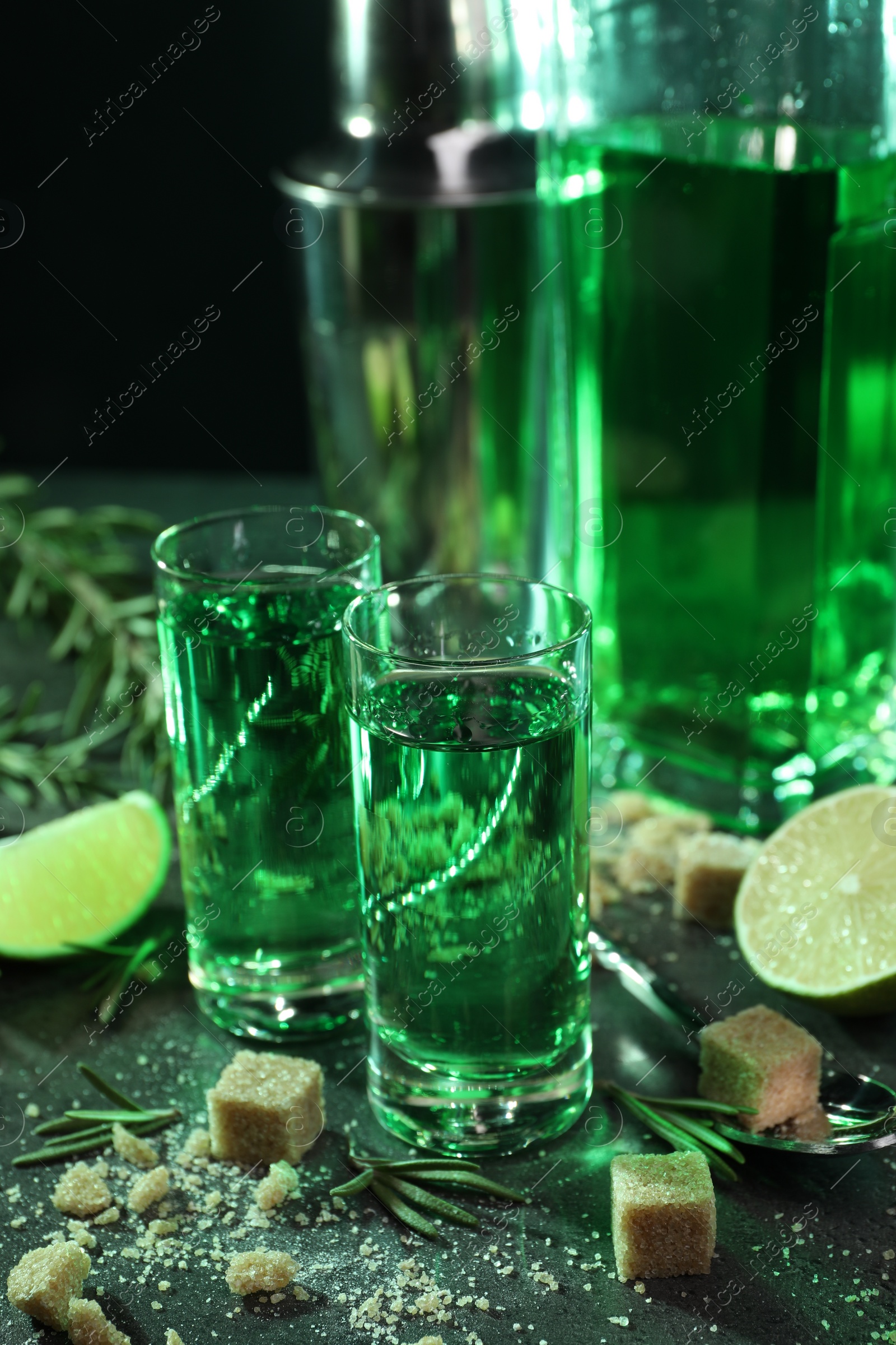 Photo of Absinthe in shot glasses, spoon, brown sugar, lime and rosemary on gray table, closeup. Alcoholic drink