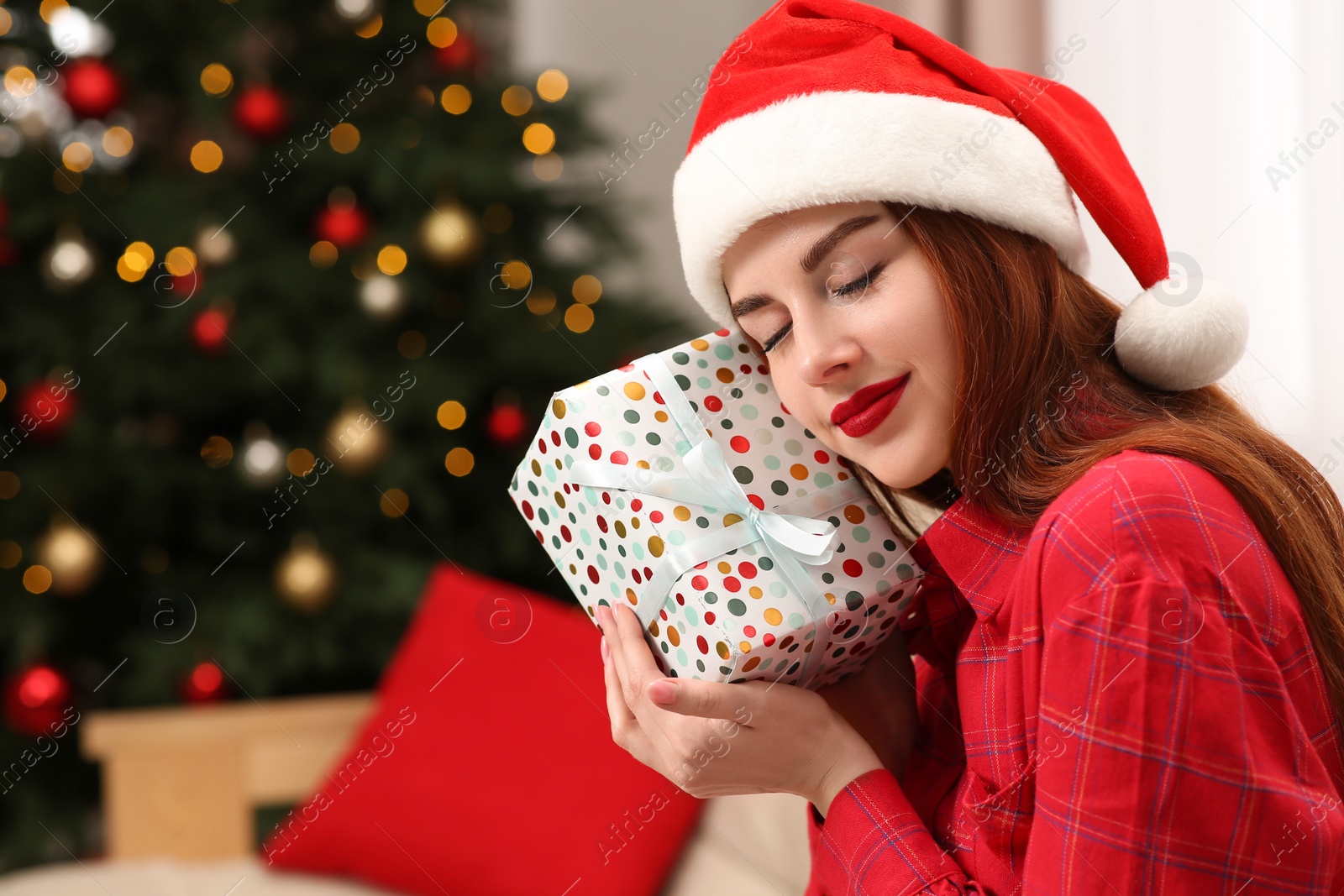 Photo of Beautiful young woman in Santa hat with gift box near Christmas tree at home, space for text