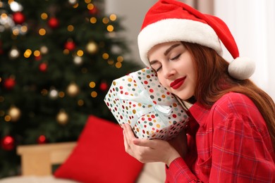 Beautiful young woman in Santa hat with gift box near Christmas tree at home, space for text