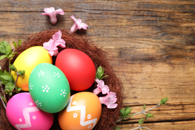 Photo of Colorful Easter eggs in decorative nest with flowers on wooden background, flat lay. Space for text