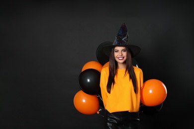 Beautiful woman wearing witch costume with balloons for Halloween party on black background, space for text