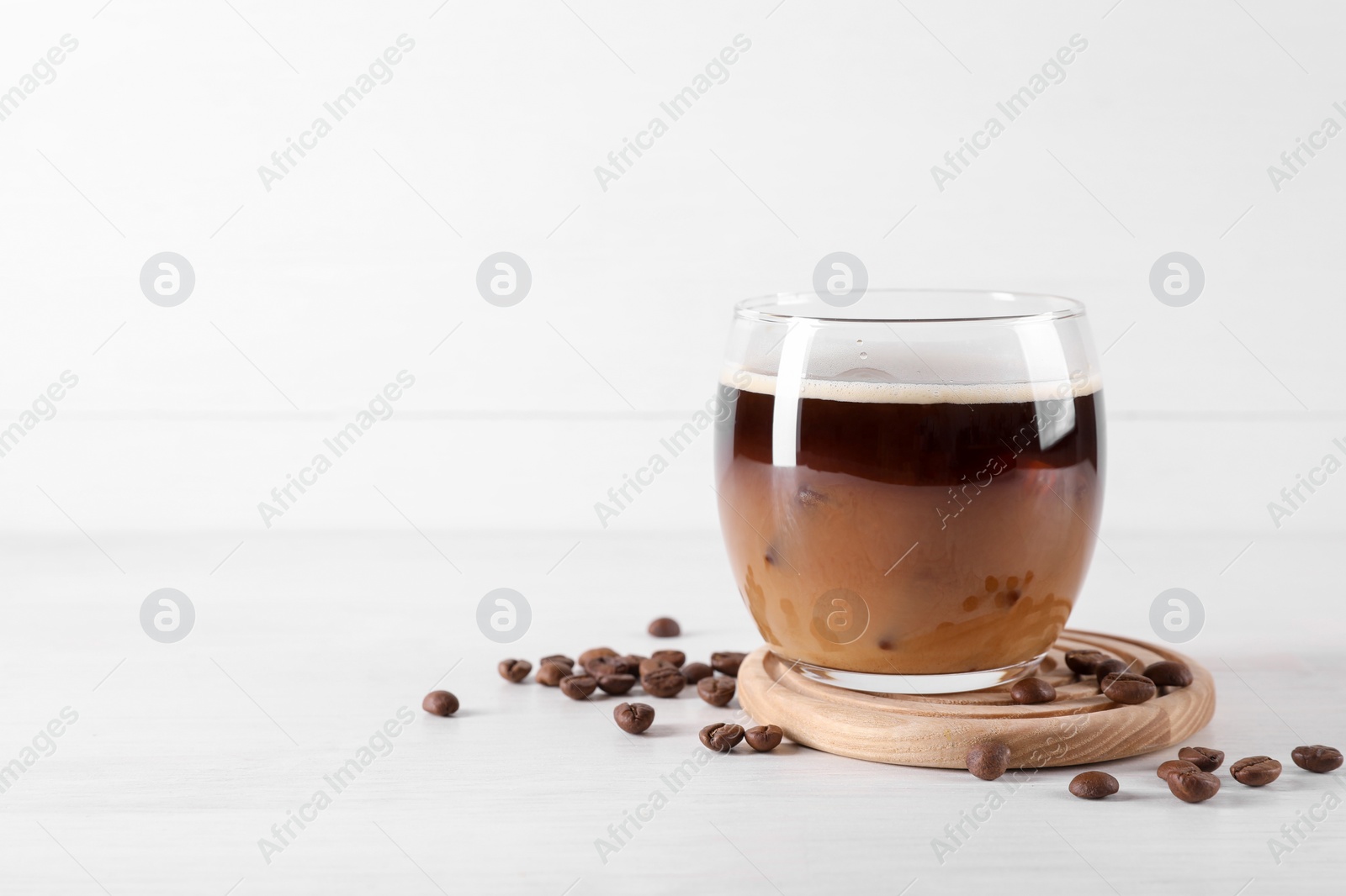 Photo of Refreshing iced coffee with milk in glass and beans on white wooden table, space for text