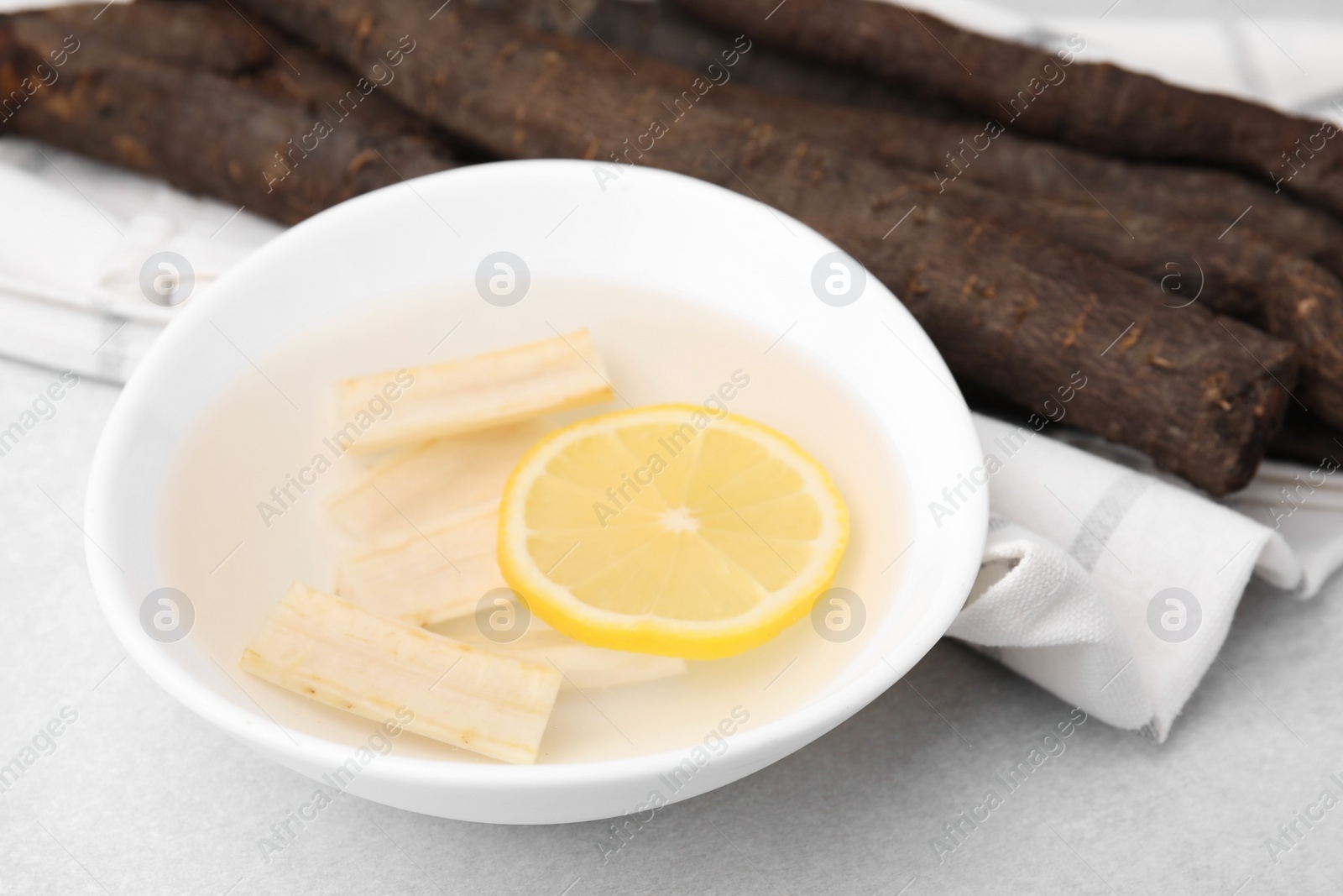 Photo of Bowl with water, lemon and raw salsify roots on white table, closeup