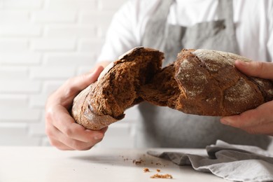 Photo of Man breaking loaf of fresh bread at white table near brick wall, closeup