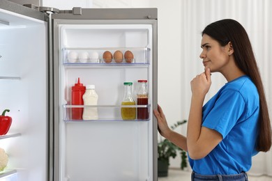 Photo of Thoughtful young woman near modern refrigerator in kitchen