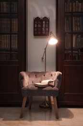 Photo of Comfortable armchair with book and lamp between wooden bookcases in library
