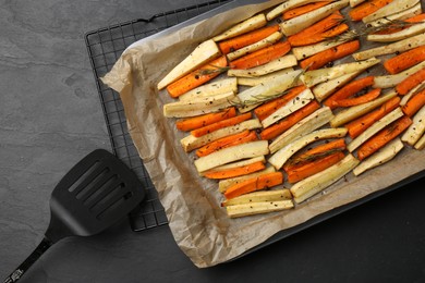 Photo of Tray with baked parsnips, carrots and spatula on black table, flat lay