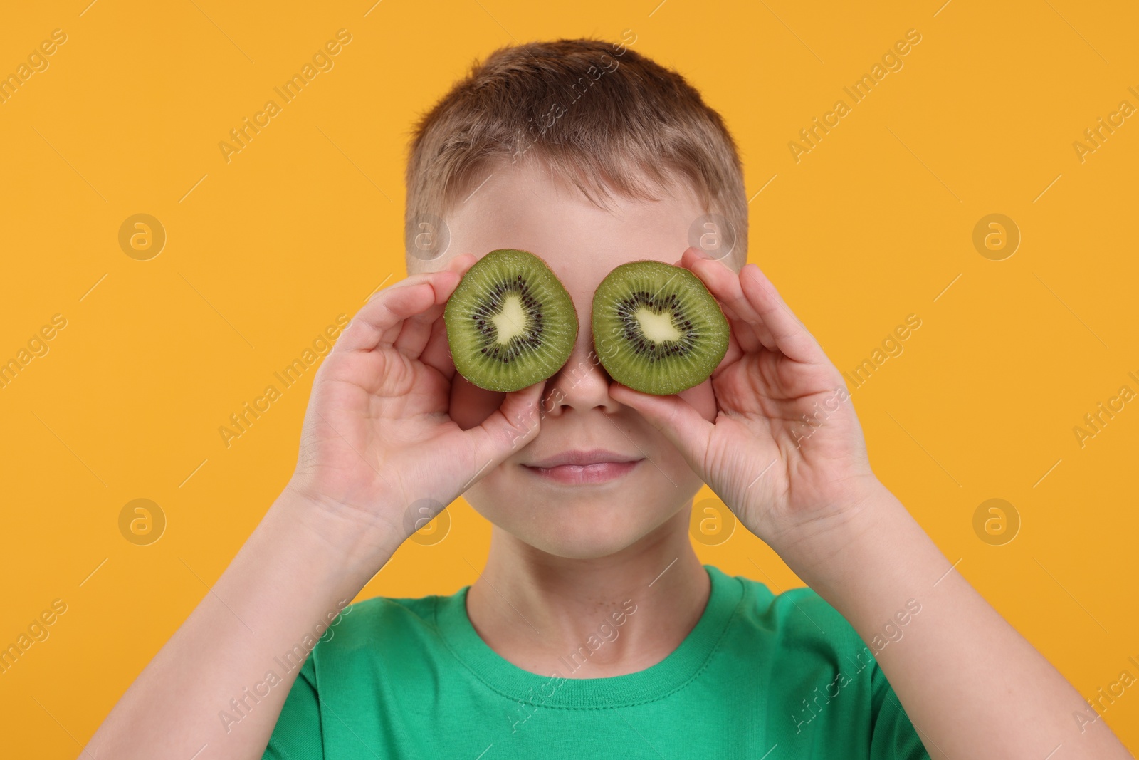 Photo of Boy covering eyes with halves of fresh kiwi on orange background