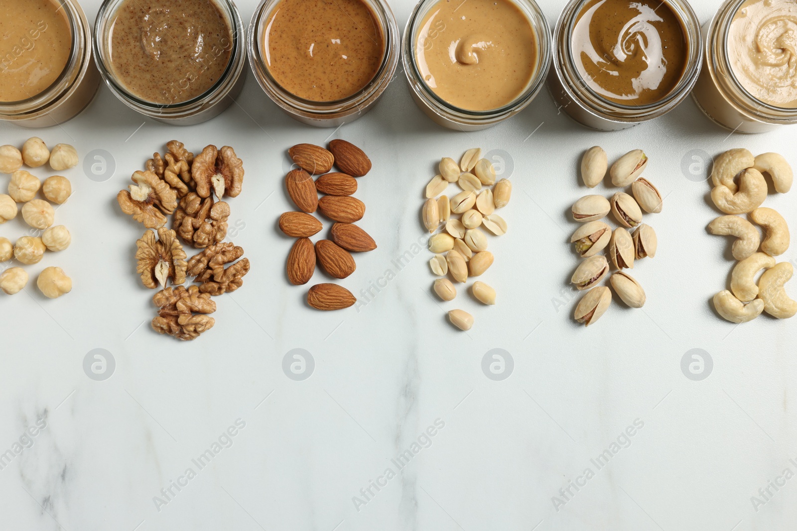 Photo of Tasty nut butters in jars and raw nuts on white marble table, flat lay. Space for text