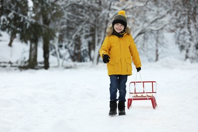Photo of Little boy pulling sledge through snow in winter park, space for text