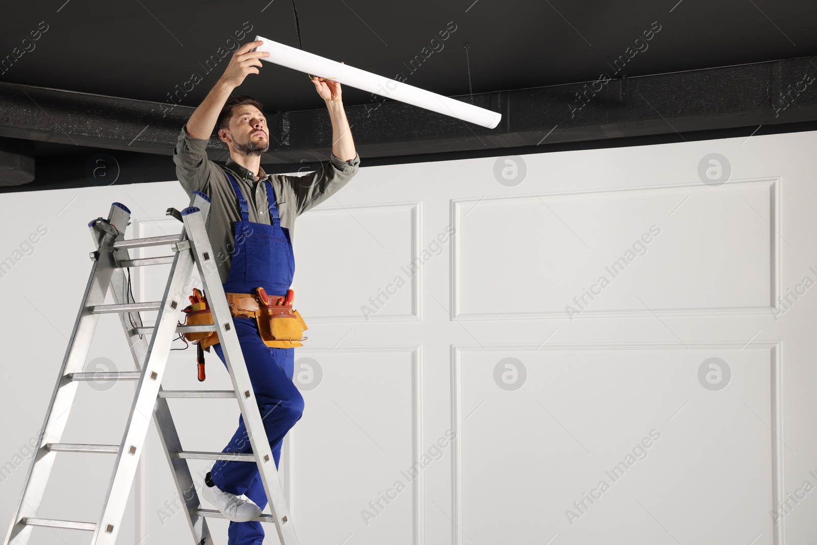 Photo of Electrician in uniform installing ceiling lamp indoors. Space for text