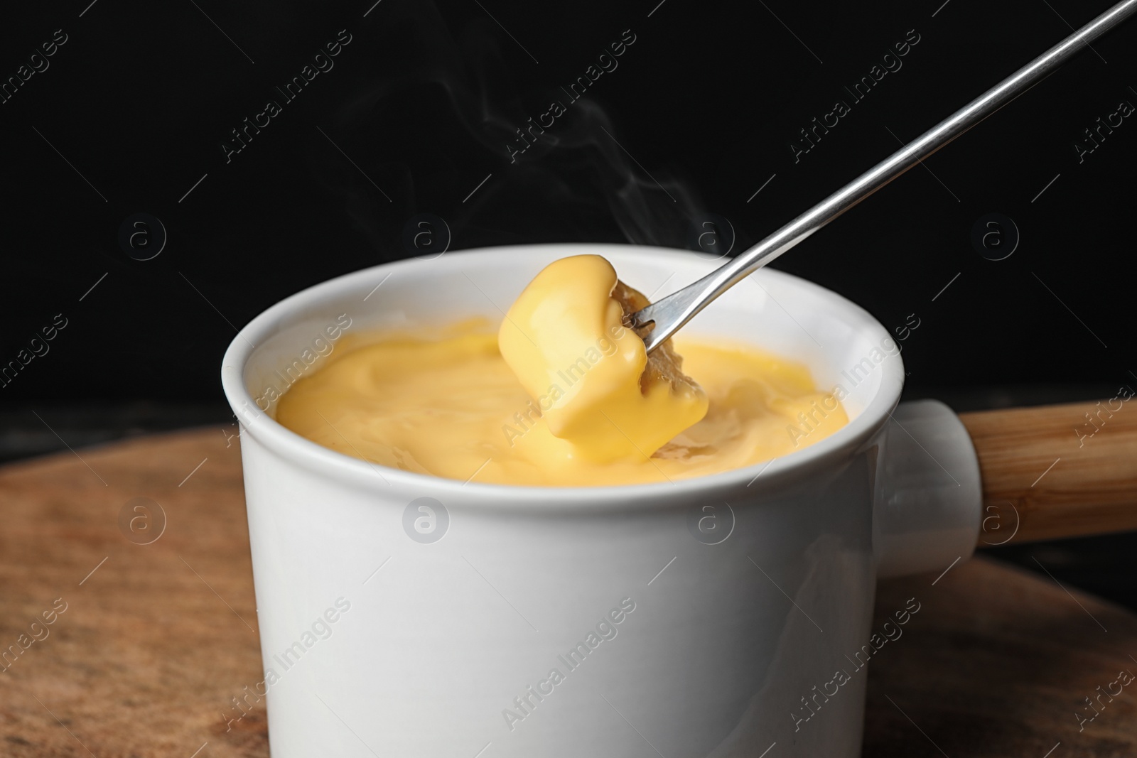 Photo of Pot of tasty cheese fondue and fork with bread on cutting board, closeup