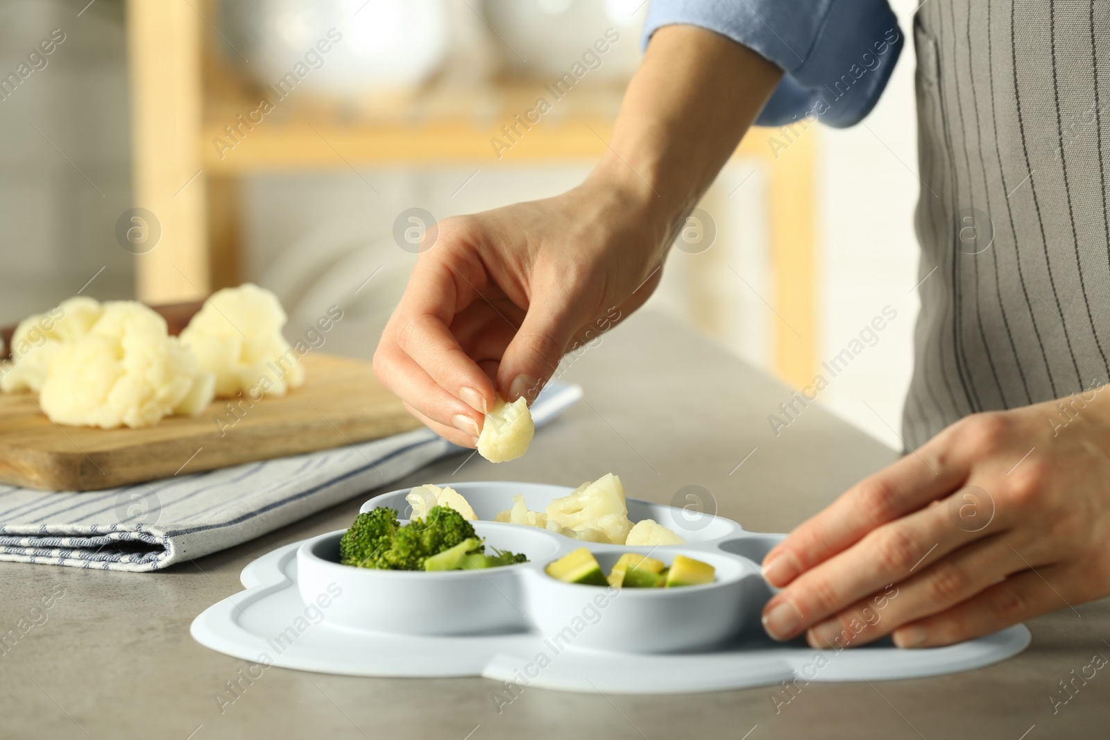 Photo of Baby food. Woman putting piece of cauliflower into section plate with different vegetables at grey textured table in kitchen, closeup