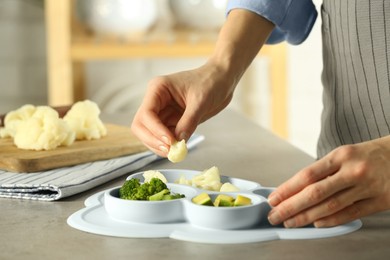Baby food. Woman putting piece of cauliflower into section plate with different vegetables at grey textured table in kitchen, closeup
