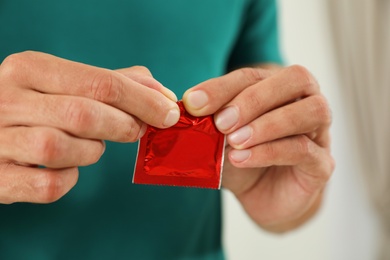 Young man opening condom on light background, closeup. Safe sex concept