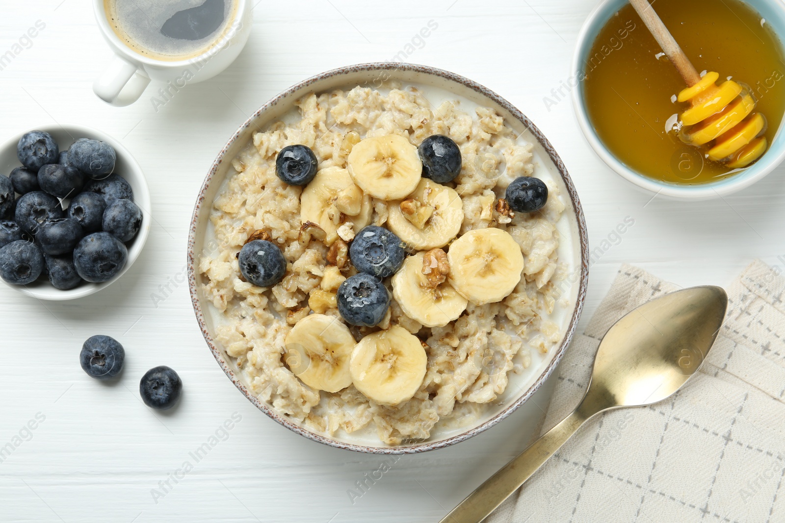 Photo of Tasty oatmeal with banana, blueberries, walnuts and honey served in bowl on white wooden table, flat lay