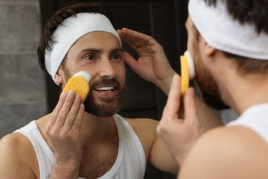 Photo of Man with headband washing his face using sponge near mirror in bathroom