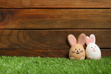 Photo of Easter eggs as cute bunnies on green grass against wooden background, space for text