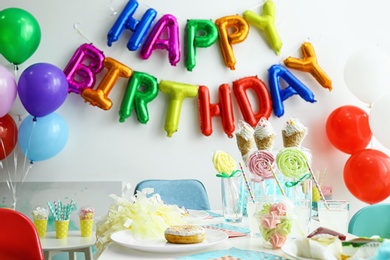 Table with treats and phrase HAPPY BIRTHDAY made of colorful balloon letters in living room