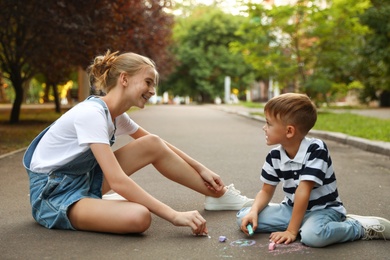 Photo of Teen nanny and cute little boy drawing with chalks on asphalt