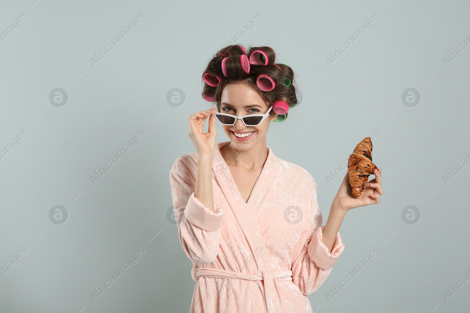 Photo of Beautiful young woman in bathrobe with hair curlers holding croissant on light grey background