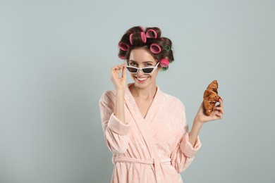 Beautiful young woman in bathrobe with hair curlers holding croissant on light grey background