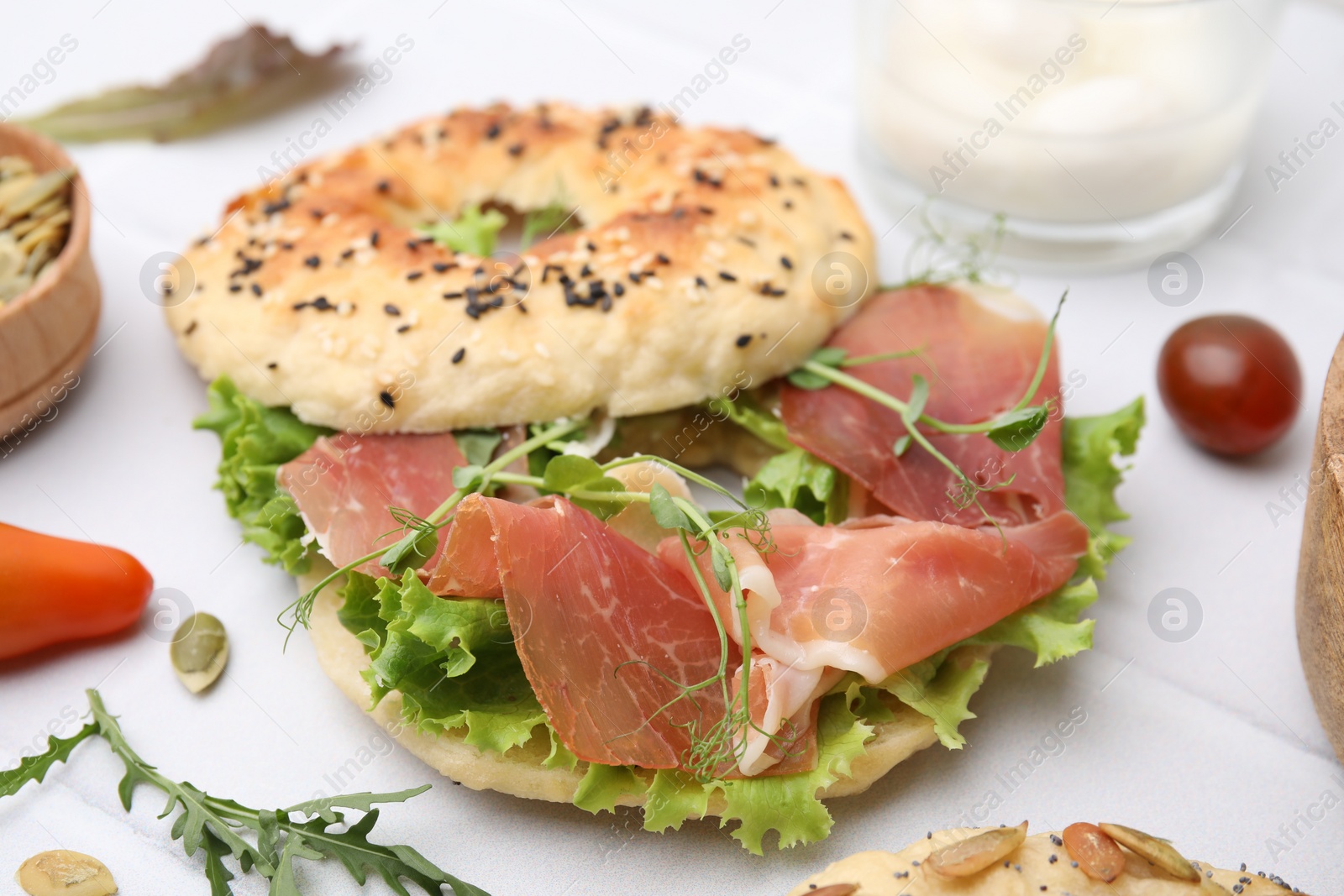 Photo of Tasty bagel with cured ham and lettuce on white tiled table, closeup