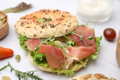 Tasty bagel with cured ham and lettuce on white tiled table, closeup