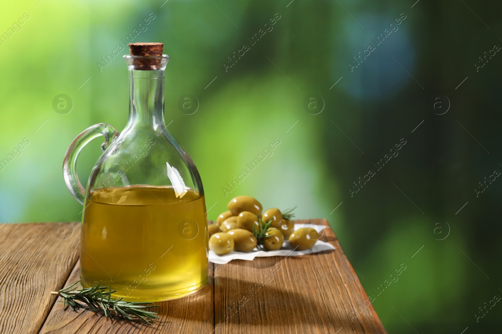 Photo of Jug of cooking oil, olives and rosemary on wooden table against blurred background. Space for text