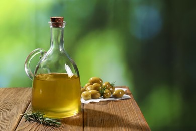 Photo of Jug of cooking oil, olives and rosemary on wooden table against blurred background. Space for text