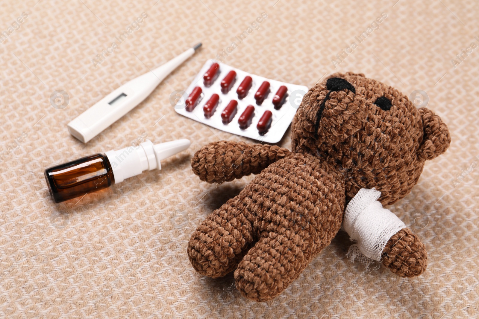 Photo of Toy bear with bandage, thermometer, pills and nasal spray on blanket, closeup