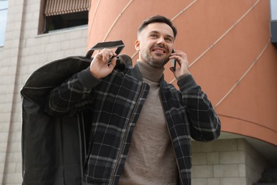 Photo of Man holding garment cover with clothes while talking on phone outdoors. Dry-cleaning service