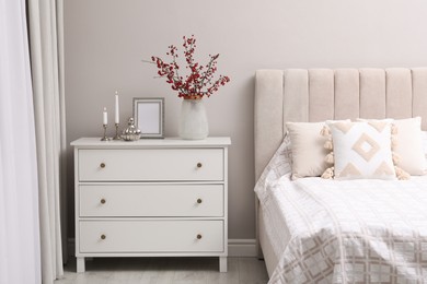 Photo of Hawthorn branches with red berries, candles and frame on chest of drawers in bedroom