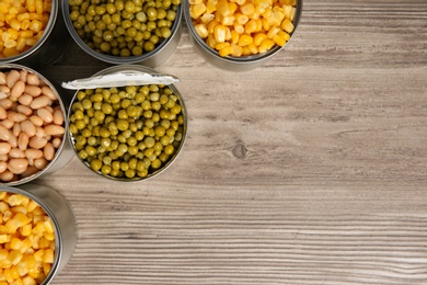Photo of Open tin cans with different vegetables on wooden table, flat lay. Space for text