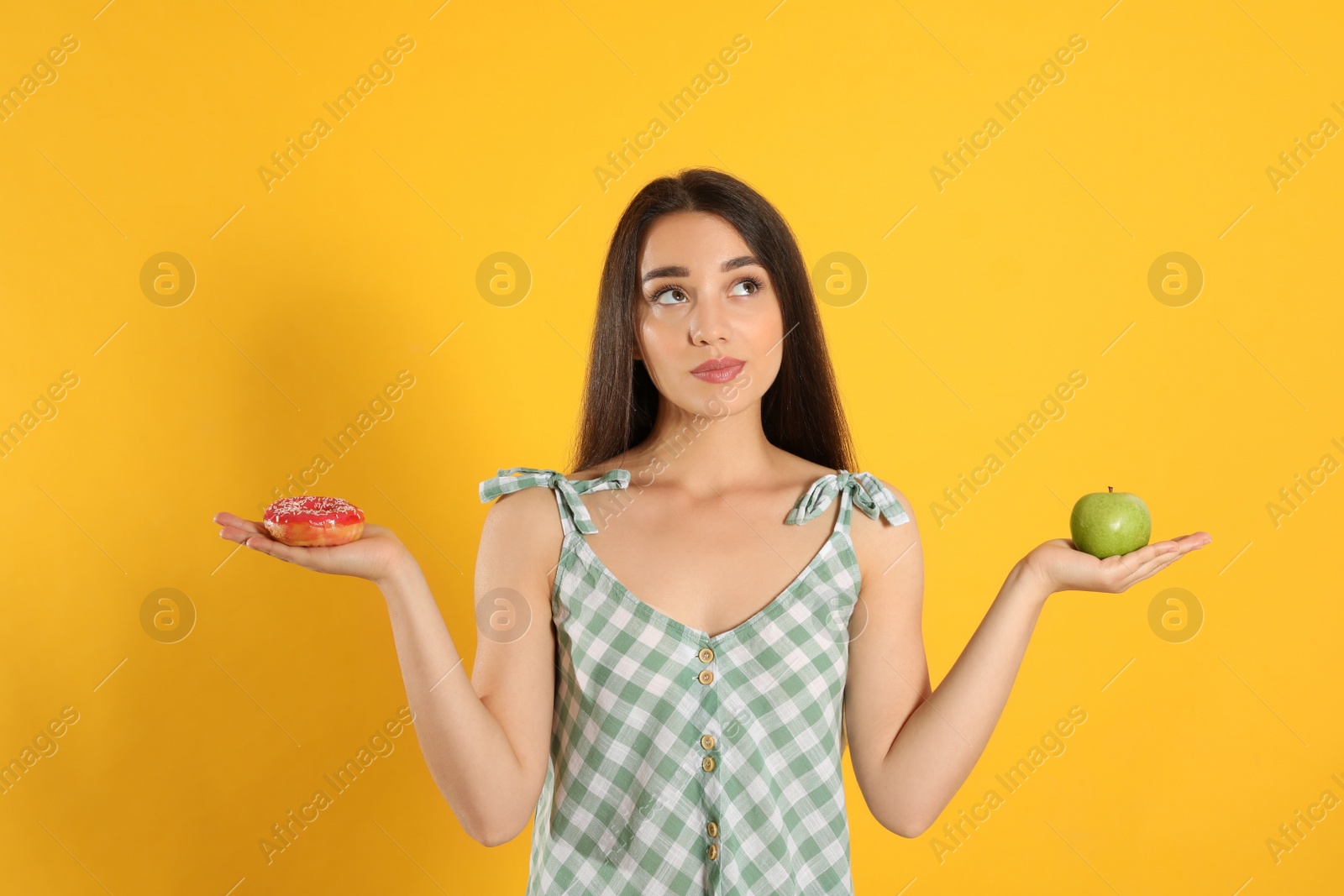 Photo of Doubtful woman choosing between apple and doughnut on yellow background