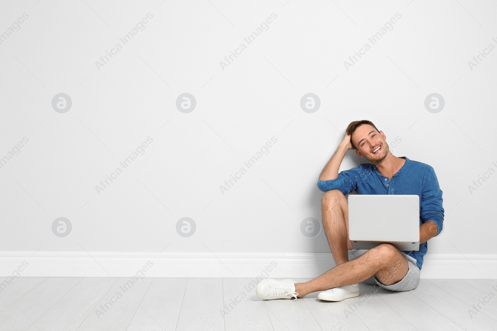 Photo of Young man with laptop sitting on floor against light wall. Space for text