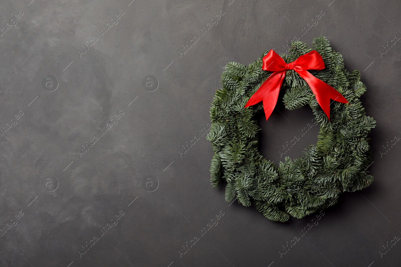 Photo of Christmas wreath made of fir tree branches with red ribbon on grey background, space for text