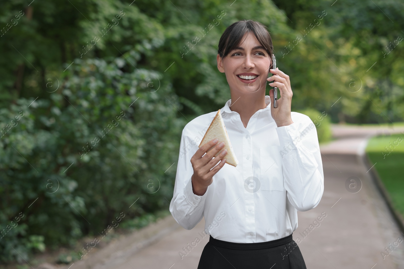 Photo of Lunch time. Happy businesswoman with sandwich talking on smartphone in park