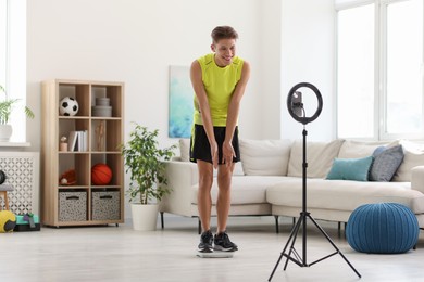 Photo of Smiling sports blogger standing on floor scales while streaming online fitness lesson with phone at home