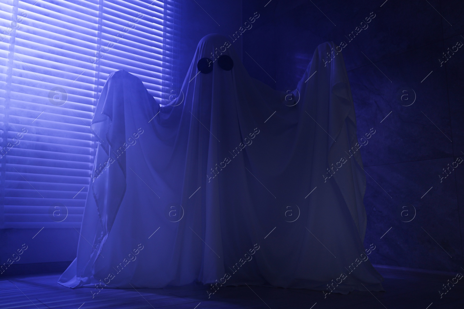 Photo of Creepy ghost. Woman covered with sheet near window in blue light