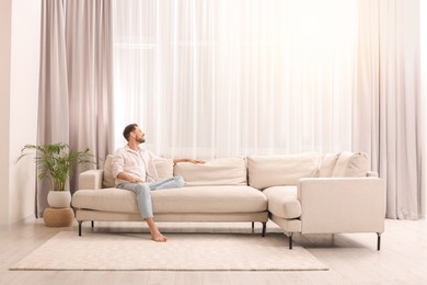 Photo of Happy man resting on sofa near window with beautiful curtains in living room