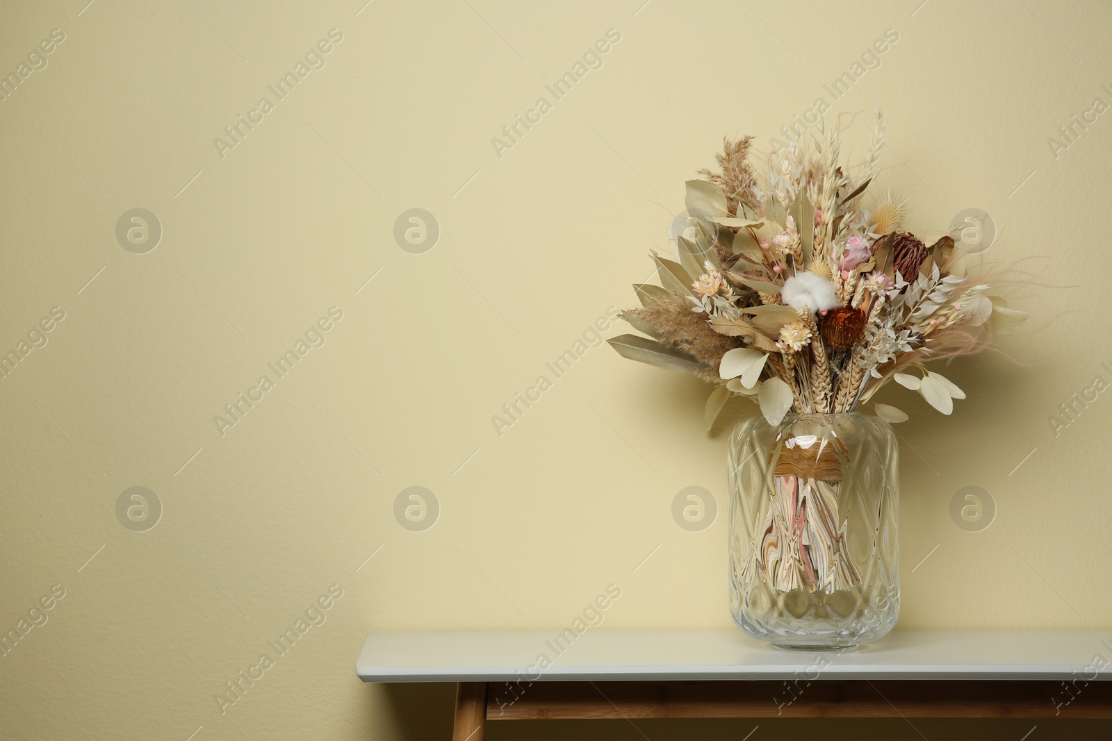 Photo of Beautiful dried flower bouquet in glass vase on white table near beige wall. Space for text