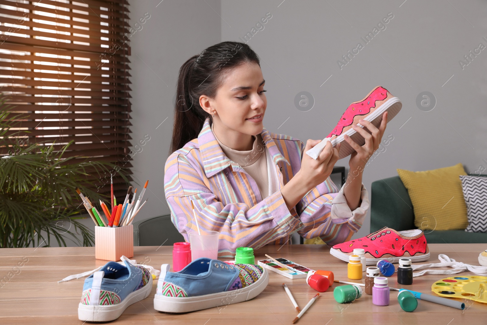 Photo of Woman painting on sneaker at wooden table indoors. Customized shoes