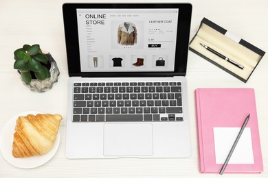 Photo of Online store. Laptop with open website, notebook, pen, croissant and pencil on wooden table, above view