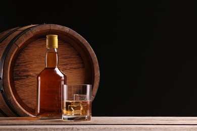 Photo of Whiskey with ice cubes in glass, bottle and barrel on wooden table against black background, space for text