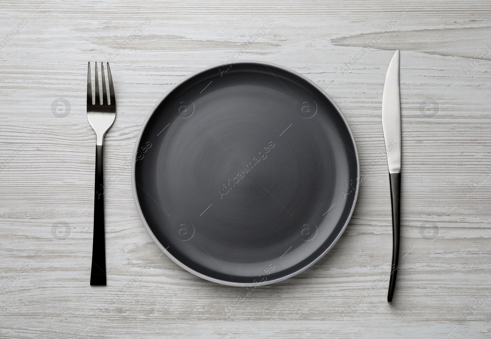 Photo of New dark plate and cutlery on light wooden table, flat lay