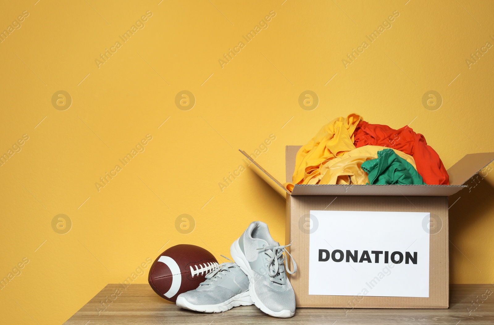 Photo of Donation box with clothes, shoes and rugby ball on table against color background. Space for text